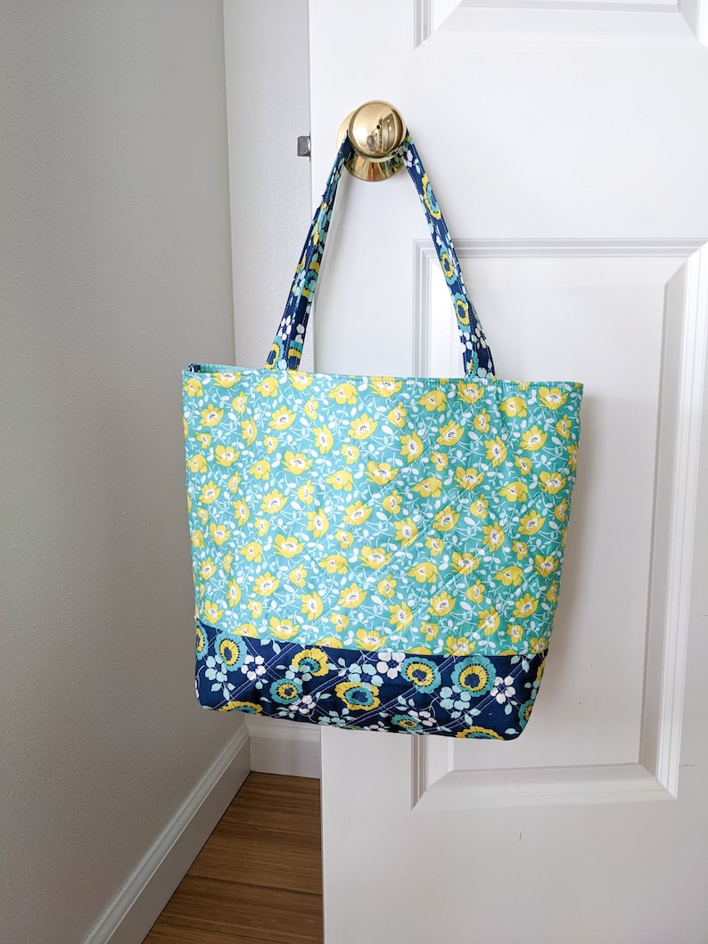 May include: A blue and green floral tote bag with yellow flowers and a navy blue trim. The bag has two straps and is hanging on a white door.