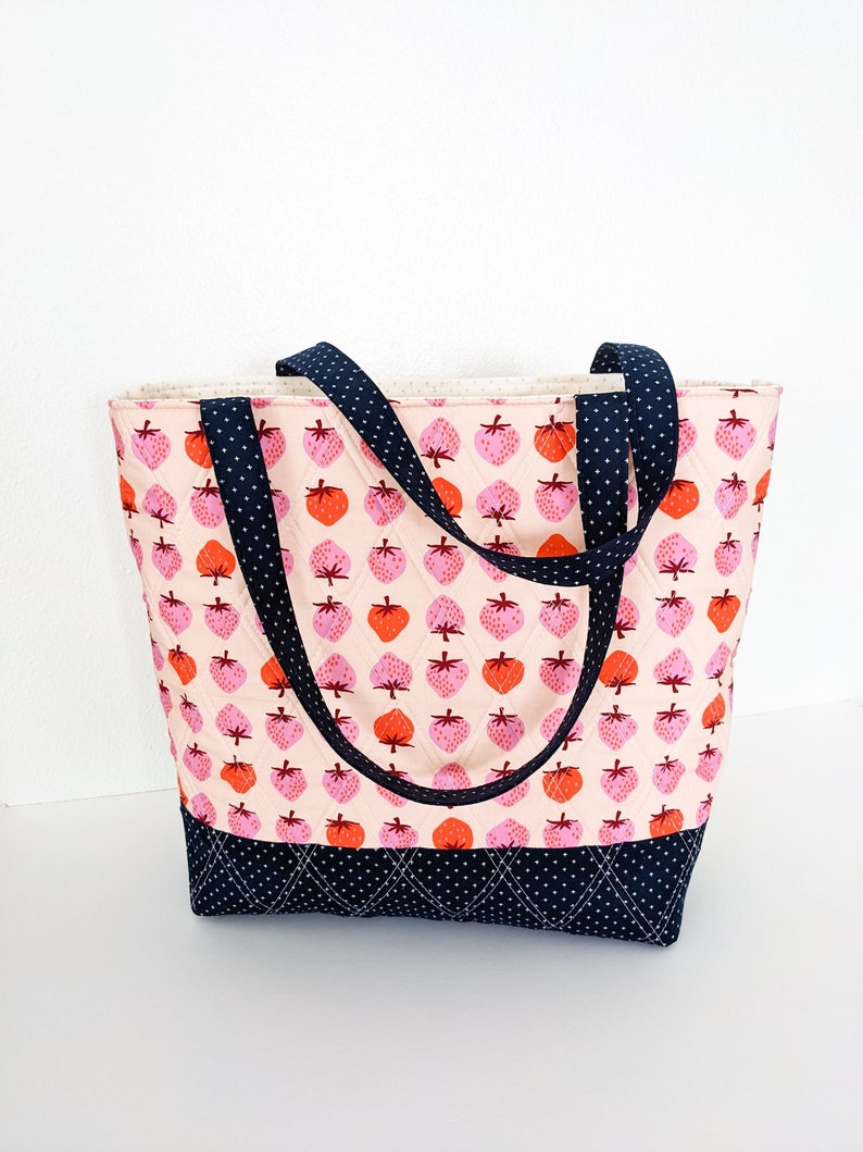 May include: A pink and white tote bag with a black and white polka dot strap. The bag is quilted and features a strawberry print.