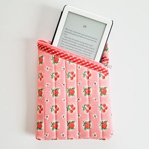 May include: A pink quilted e-reader case with a white zipper and a red and white checkered lining. The case has a pattern of red cherries and white flowers on a pink background. The case is holding a white e-reader with a black screen displaying text.
