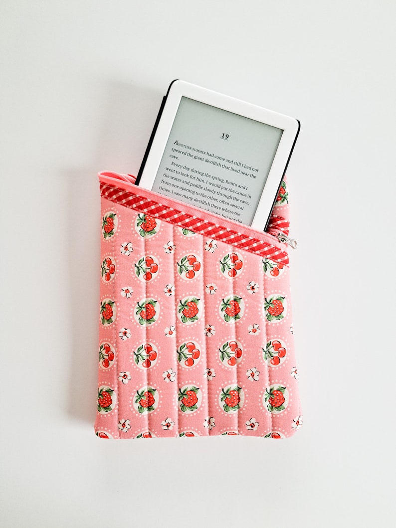 May include: A pink quilted e-reader case with a white zipper and a red and white checkered lining. The case has a pattern of red cherries and white flowers on a pink background. The case is holding a white e-reader with a black screen displaying text.