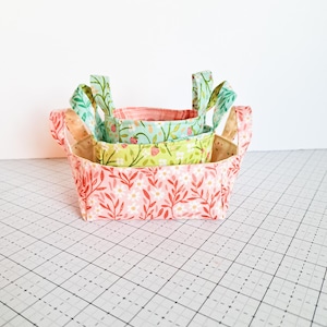 以下が含まれることがあります： 取っ手付きの布製の収納バスケットが3つ。一番大きいバスケットはピンク地に花柄、真ん中のバスケットは緑地にイチゴ柄、一番小さいバスケットは薄い青地にイチゴ柄です。