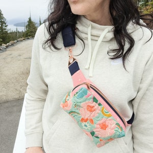 May include: A floral patterned fanny pack with a navy blue adjustable strap and rose gold hardware. The fanny pack is worn by a person wearing a white hoodie.