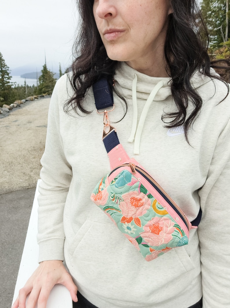 May include: A floral patterned fanny pack with a navy blue adjustable strap and rose gold hardware. The fanny pack is worn by a person wearing a white hoodie.