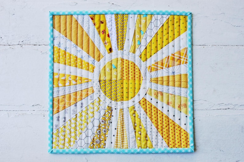May include: A yellow and white quilted sunburst design with a blue and white gingham border. The sunburst is made of various yellow and white fabrics with different patterns.