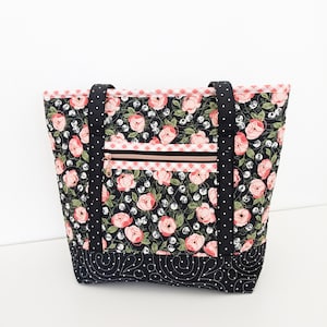 Op de afbeelding: Een zwarte quilted tote bag met een roze bloemenprint en zwarte stippen. De tas heeft twee zwarte hengsels en een ritsvak met een gouden rits.