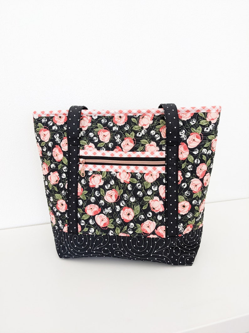 Op de afbeelding: Een zwarte quilted tote bag met een roze bloemenprint en zwarte stippen. De tas heeft twee zwarte hengsels en een ritsvak met een gouden rits.