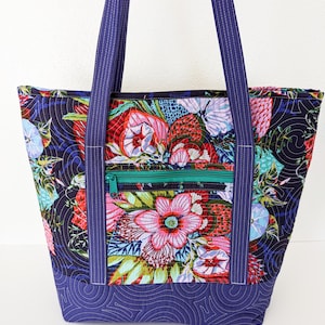 Op de afbeelding: Een quilted tote bag met een bloemenprint in tinten roze, rood, groen en blauw. De tas heeft een ritsvak en donkerblauwe hengsels.