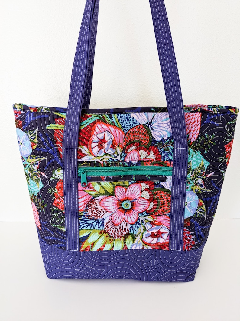 Op de afbeelding: Een quilted tote bag met een bloemenprint in tinten roze, rood, groen en blauw. De tas heeft een ritsvak en donkerblauwe hengsels.
