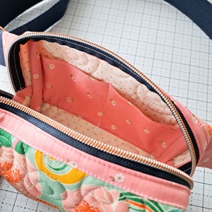 May include: A pink quilted fanny pack with a floral print and a black zipper. The inside of the bag is lined with a pink fabric with a white floral pattern.