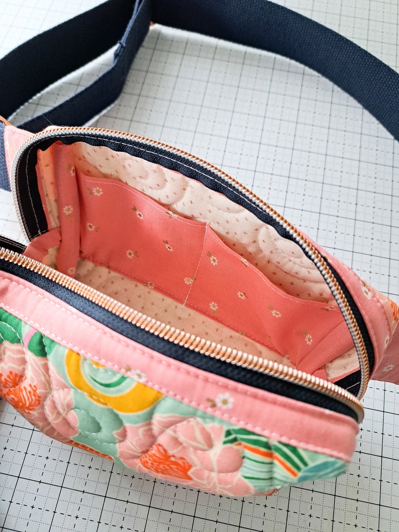 May include: A pink quilted fanny pack with a floral print and a black zipper. The inside of the bag is lined with a pink fabric with a white floral pattern.
