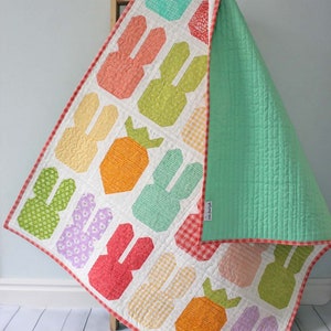May include: A white baby quilt with a patchwork design featuring carrots and bunnies. The quilt is made with a variety of colorful fabrics, including orange, green, pink, and yellow. The quilt is backed with a solid green fabric and has a red and white gingham border.