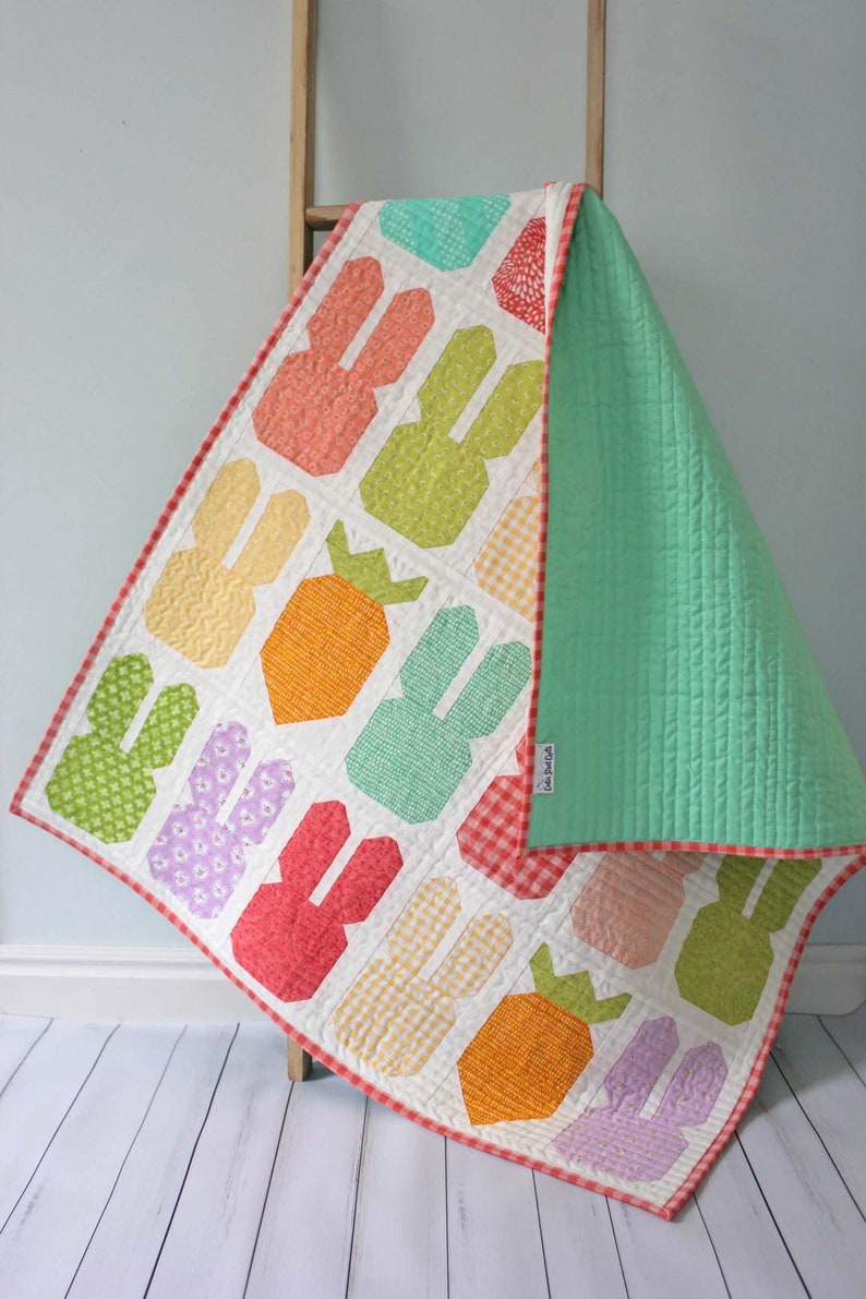 May include: A white baby quilt with a patchwork design featuring carrots and bunnies. The quilt is made with a variety of colorful fabrics, including orange, green, pink, and yellow. The quilt is backed with a solid green fabric and has a red and white gingham border.