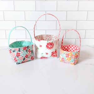 May include: Three fabric baskets with handles. The baskets are lined with different floral patterns. The largest basket is white with red roses, the medium basket is blue with pink and white flowers, and the smallest basket is red and white gingham with orange and pink flowers.