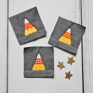 May include: Three gray fabric squares with a candy corn design in orange, yellow, and white fabric. Three wooden star-shaped buttons are scattered on a white wooden surface.