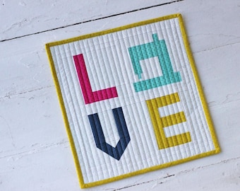 May include: A small quilted wall hanging with the word "LOVE" spelled out in colorful fabric letters. The letters are outlined in yellow and the background is white with a subtle polka dot pattern.