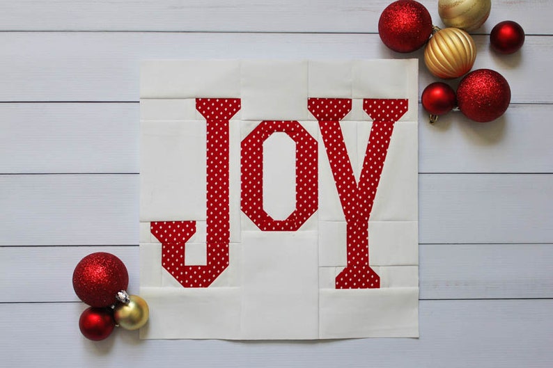 May include: A white fabric square with the word "JOY" in red with white polka dots. The fabric is quilted and the letters are appliqued. The background is a white wood surface with red and gold ornaments.