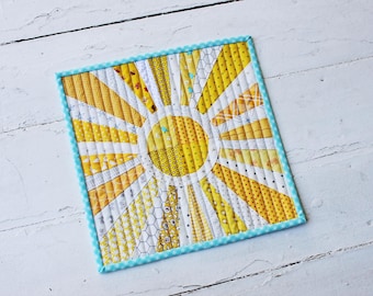 Puede incluir: Un diseño de sol radiante acolchado en amarillo y blanco con un borde a cuadros azul y blanco. El sol radiante está hecho de recortes de tela en varios patrones amarillos y blancos.