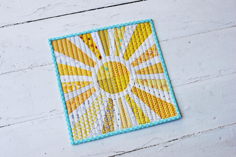 May include: A yellow and white quilted sunburst design with a blue and white checkered border. The sunburst is made of fabric scraps in various yellow and white patterns.