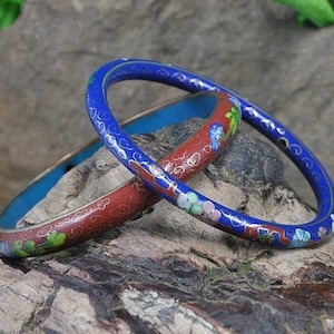 May include: Two enamel bangles, one blue and one red, with floral designs. The bangles are stacked on a piece of driftwood.