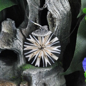 May include: A black and white round pendant with a silver chain. The pendant has a starburst design with a white center and black rays. The pendant is sitting on a piece of driftwood with green leaves and purple flowers in the background.