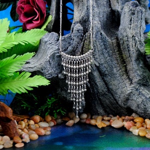 May include: A silver necklace with a cascading design of small beads. The necklace is displayed on a wooden surface with green foliage and small stones.