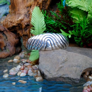 May include: A silver bracelet with a wavy, textured design. The bracelet is resting on a gray rock.