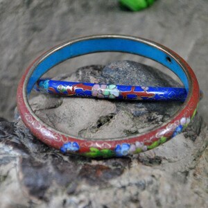 May include: Two enamel bangles, one red with floral designs and one blue with white flowers, on a gray rock background.
