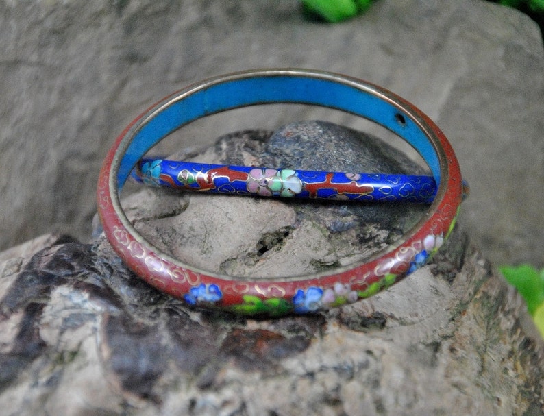 May include: Two enamel bangles, one red with floral designs and one blue with white flowers, on a gray rock background.
