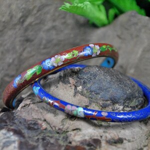 May include: Two enamel bracelets with floral designs. The top bracelet is red with blue and white flowers. The bottom bracelet is blue with red and white flowers.
