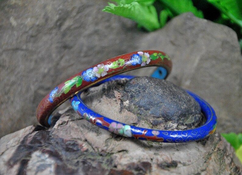 May include: Two enamel bracelets with floral designs. The top bracelet is red with blue and white flowers. The bottom bracelet is blue with red and white flowers.