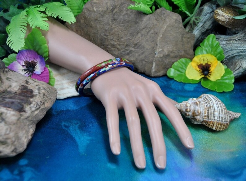 May include: Two colorful bangle bracelets with floral patterns in shades of blue, red, and white. The bracelets are stacked on a mannequin's wrist.