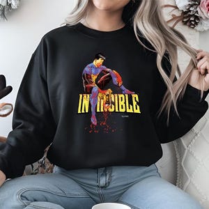 May include: Black sweatshirt featuring a colorful graphic of a superhero with the word "INVINCIBLE" in yellow block letters. The superhero is depicted in a seated position with a red and blue color scheme. The sweatshirt is a casual style.