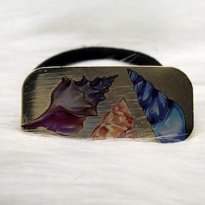 May include: A gold-colored hair tie with a rectangular metal charm featuring three seashells. The seashells are painted in shades of purple, blue, and pink.