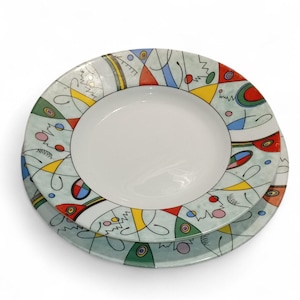 May include: Two ceramic plates, one stacked on the other. The plates have a white center and a light blue rim with colorful abstract designs in red, yellow, blue, and green. The plates are on a white background.