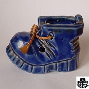 Può includere: Un piccolo stivale blu in ceramica con un laccio dorato. Lo stivale ha una finitura lucida ed è dettagliato con creste e fori. Lo stivale è un oggetto decorativo.