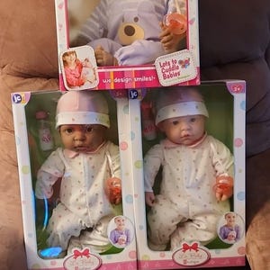 May include: Three baby dolls in boxes. The dolls have soft skin and wear pajamas and hats. The boxes have the text "Lots to Cuddle Babies" and "La Baby Boutique". One doll wears a purple outfit, and the other two wear white outfits.