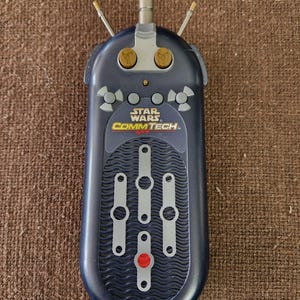 May include: A dark blue Star Wars CommTech device with silver and gold accents. The device has a series of gray sliders and a red button. The Star Wars CommTech logo is visible on the front.