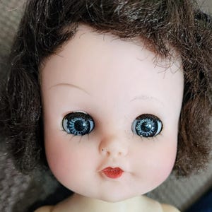 May include: A doll with short, wavy brown hair, pale skin, and large, round blue eyes with dark lashes. The doll features a small, painted red mouth, a rounded nose, and subtle pink blush on the cheeks, capturing a detailed facial expression.