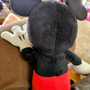 Peut inclure: Une peluche Mickey Mouse, vue de dos, pr&eacute;sentant ses c&eacute;l&egrave;bres oreilles noires, son corps noir, son short rouge et ses gants blancs. Le jouet en peluche est pos&eacute; sur une surface marron, avec les d&eacute;tails de son design clairement visibles.