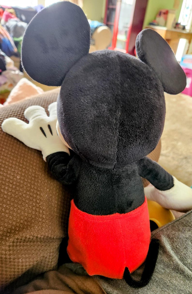 Peut inclure: Une peluche Mickey Mouse, vue de dos, pr&eacute;sentant ses c&eacute;l&egrave;bres oreilles noires, son corps noir, son short rouge et ses gants blancs. Le jouet en peluche est pos&eacute; sur une surface marron, avec les d&eacute;tails de son design clairement visibles.