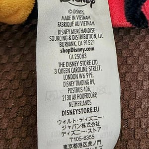 Peut inclure: Gros plan sur une &eacute;tiquette blanche avec le logo Disney et du texte, dont "MADE IN VIETNAM" et "shopDisney.com". L'&eacute;tiquette est partiellement visible sur un fond de tissu rouge, noir et jaune. L'&eacute;tiquette contient &eacute;galement l'adresse de The Disney Store Ltd &agrave; Londres.