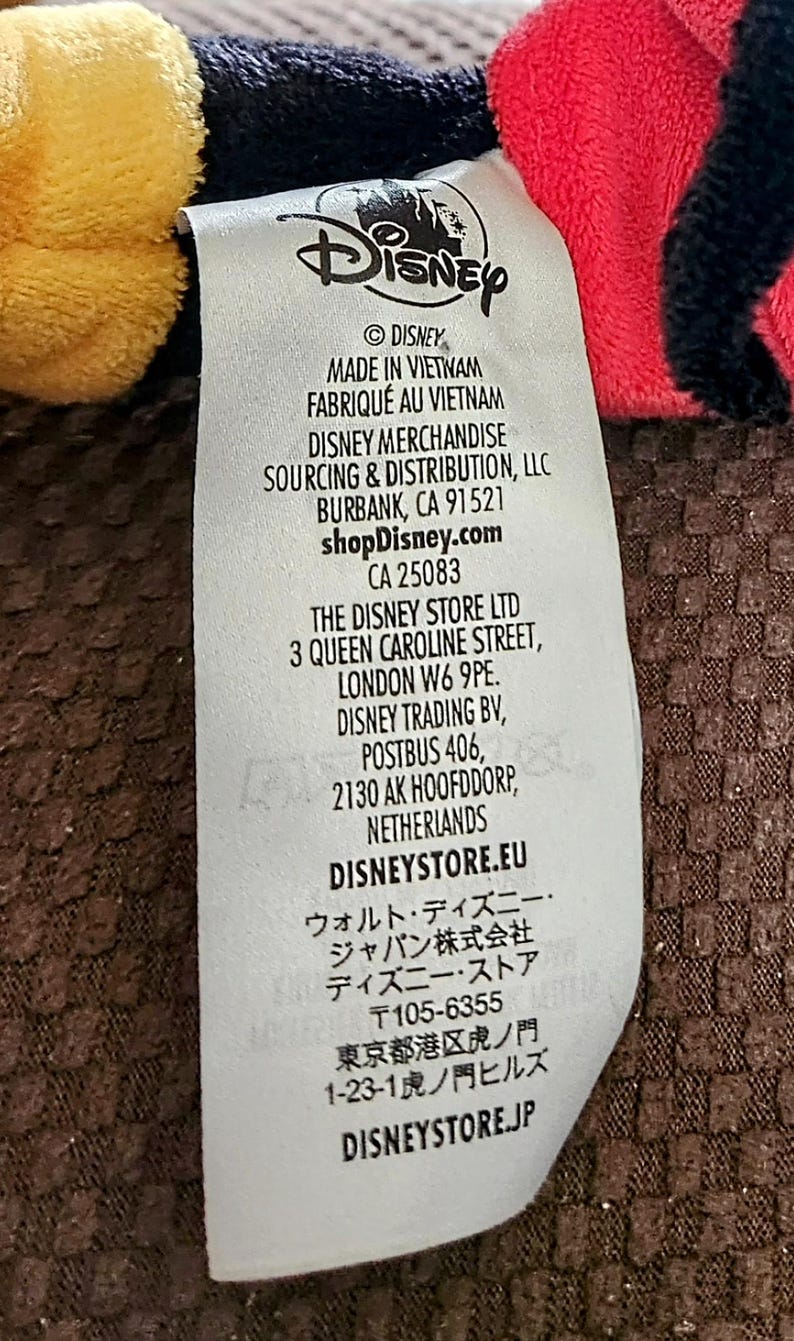 Peut inclure: Gros plan sur une &eacute;tiquette blanche avec le logo Disney et du texte, dont "MADE IN VIETNAM" et "shopDisney.com". L'&eacute;tiquette est partiellement visible sur un fond de tissu rouge, noir et jaune. L'&eacute;tiquette contient &eacute;galement l'adresse de The Disney Store Ltd &agrave; Londres.