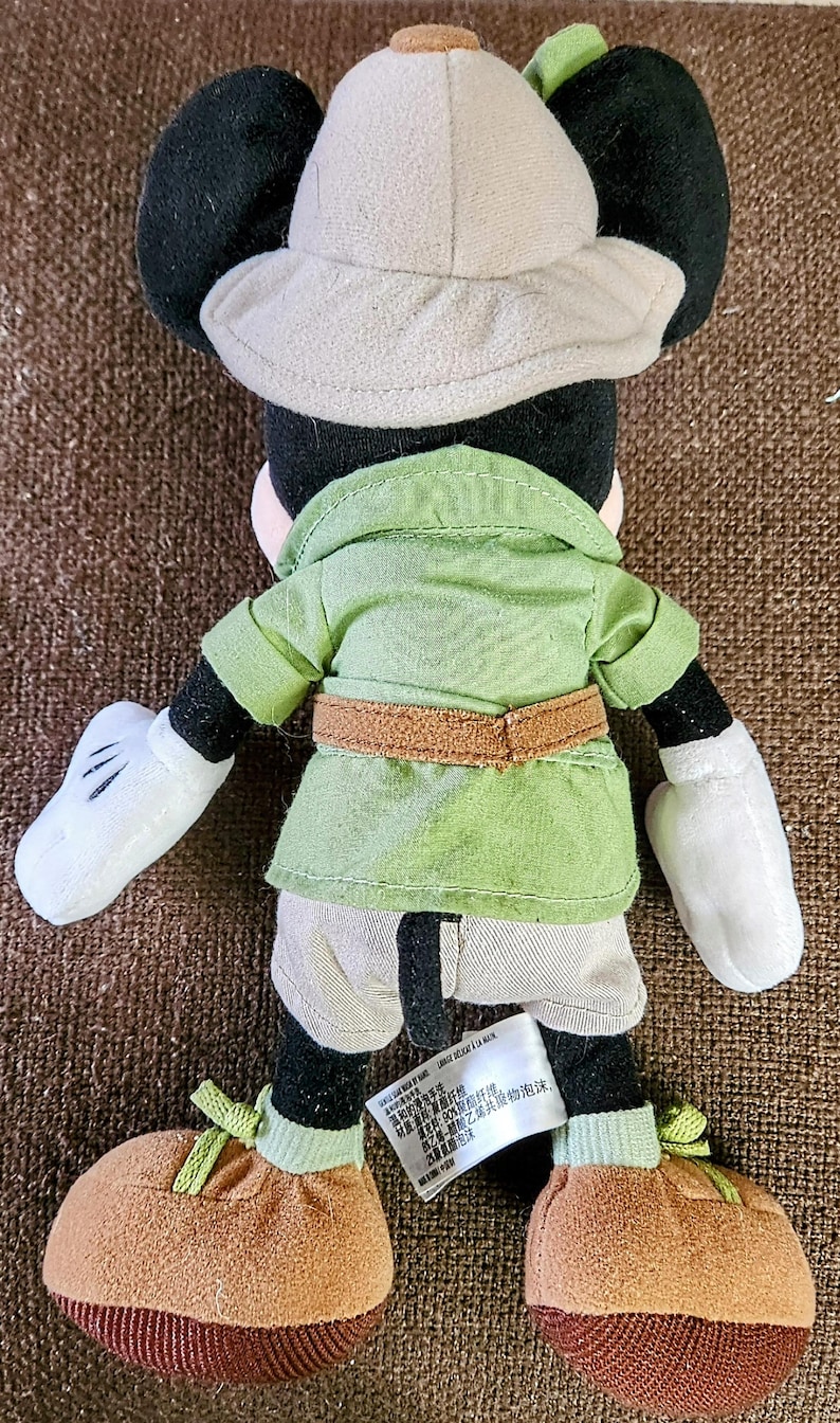 Puede incluir: Un peluche de Mickey Mouse con un atuendo de safari. El juguete tiene orejas negras, un sombrero de safari beige, una camisa verde con cintur&oacute;n marr&oacute;n y pantalones cortos color caqui. Los zapatos son marrones con cordones verdes.
