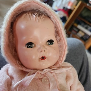 May include: A vintage baby doll with a pink hooded coat. The doll has a painted face with blue eyes and red lips. The coat has a button closure and ribbon ties. The doll's skin has a slightly aged appearance.