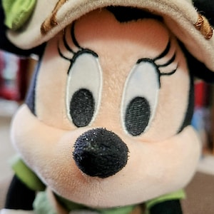 Puede incluir: Mu&ntilde;eca de peluche de Minnie Mouse con un sombrero de estilo safari con un lazo verde y un atuendo verde y blanco. La mu&ntilde;eca tiene ojos grandes y expresivos, una nariz negra y orejas negras. Es un objeto de colecci&oacute;n.