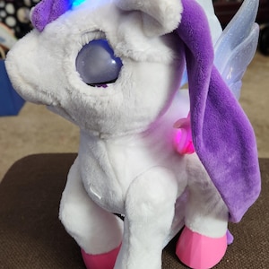 May include: A plush unicorn toy with white fur, a glowing blue horn, and purple fabric accents. It features large, dark, glossy eyes, translucent glittery wings, and bright pink, faceted hooves. The toy has an integrated light-up mechanism.