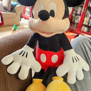 Peut inclure: Poup&eacute;e en peluche de Mickey Mouse avec des oreilles noires, un visage beige et un nez noir. La poup&eacute;e porte une chemise noire, un short rouge avec des boutons blancs, des gants blancs et des chaussures jaunes. La poup&eacute;e est assise sur une surface marron.