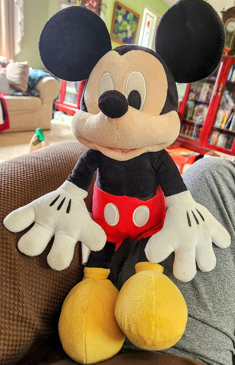 Peut inclure: Poup&eacute;e en peluche de Mickey Mouse avec des oreilles noires, un visage beige et un nez noir. La poup&eacute;e porte une chemise noire, un short rouge avec des boutons blancs, des gants blancs et des chaussures jaunes. La poup&eacute;e est assise sur une surface marron.