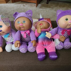 May include: Four Cabbage Patch Kids dolls in plush costumes. The first is a dog with blue eyes and heart-patterned ears. The second is a cat with green eyes and a pink bow. The third is a unicorn with a horn. The fourth is a cat with green eyes.