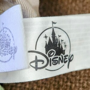 Puede incluir: Primer plano de una etiqueta de tela blanca con el logotipo de Disney y una silueta negra de un castillo. El logotipo de Disney est&aacute; en una fuente cursiva. El castillo tiene m&uacute;ltiples torres y una bandera.